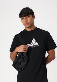Sort bomulds T-shirt med en grafisk design af en lastbil og en trekant. Modellen holder en sort stoftaske med en tekstureret overflade.