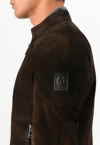 Jachetă din suede maro cu guler înalt, închidere cu fermoar și un material texturat. Prezintă o insignă neagră pe partea superioară a brațului.
