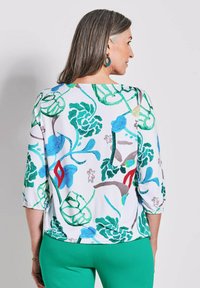 Multicolore bloes met bloemenmotief en driekwartmouwen. Heeft een witte achtergrond, groene en blauwe ontwerpen, en elastische manchetten.