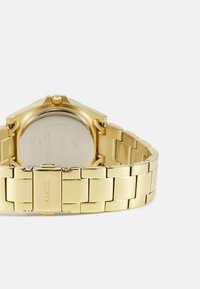 Montre en métal doré avec un bracelet à maillons, présentant un cadran rond et un dos en acier inoxydable. Des gravures sont visibles à l'arrière.