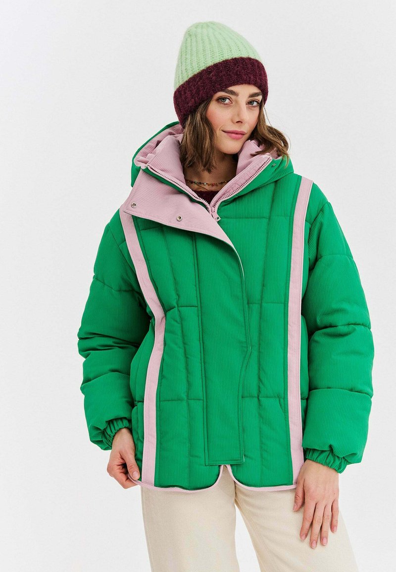 Zelená prešívaná bunda s ružovými akcentmi, ktorá má vysoký golier, zapínanie na cvoky a rebrovanú textúru. Modelka má na sebe zelenú a bordovú čiapku.