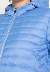 Doudoune bleu clair avec capuche, présentant des coutures matelassées verticales et une fermeture éclair argentée. Texture douce, design léger.