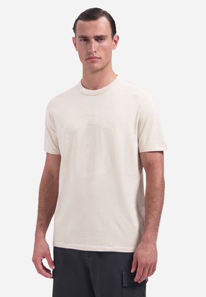 Mann trägt ein schlichtes hellbeiges T-Shirt mit kurzen Ärmeln und dunkle Cargohosen, steht vor einem einfarbigen weißen Hintergrund.