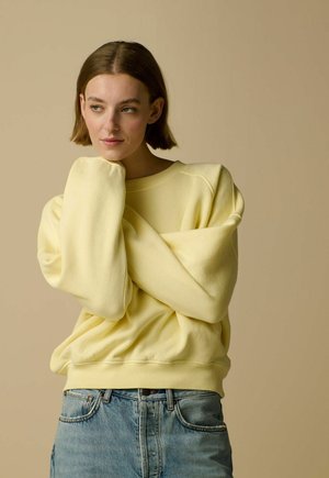 Woman with short brown hair wearing a light yellow oversized sweatshirt and blue jeans, resting her head on her hand against a beige background.