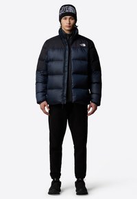 Vadderad marinblå jacka med svarta detaljer, hög krage och dragkedja framtill. Bärs med svarta byxor och en matchande beanie med logodesign.