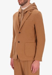 Blazer marrone realizzato in tessuto morbido con revers a punta, due tasche frontali e due bottoni. Indossato sopra un hoodie beige per un look a strati.