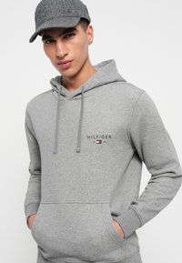 Young man wearing grey Tommy Hilfiger hoodie and matching grey cap, hands in hoodie pocket, against plain light background.