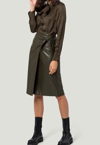 Blouse en satin vert olive avec détail à nouer, associée à une jupe en cuir synthétique vert olive de longueur genou avec un design en nœud torsadé. Des bottines noires à chevilles complètent le look.