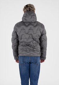 Grijze pufferjack met een capuchon, met een gewatteerd,_TEXTUURED ontwerp. Gecombineerd met blauwe spijkerbroeken. Zicht van achteren.