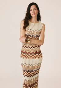 Femme aux longs cheveux foncés, debout, portant une robe beige sans manches avec des rayures ondulées marron, accessoirisée avec des bracelets épais à son poignet gauche.