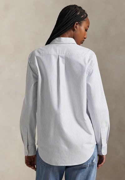 Kvinde med fletninger iført en hvid skjorte med tynde lodrette grå striber og blå jeans, set bagfra mod en neutral baggrund.