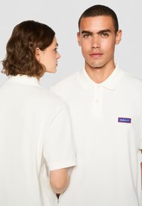 Valge polo särk pehme tekstuuriga, klassikalise kraega, kahe nupuga ja ristküliku kujulise punase ja sinise logoga rinnal.
