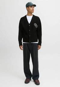 Schwarze gestrickte Strickjacke mit Knopfleiste, ausgestattet mit einem gestickten Logo. Kombiniert mit vertikal gestreiften Hosen und einer grünen Kappe.