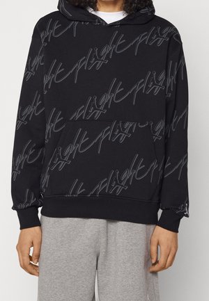 Czarna bluza z kapturem z szarym napisem "flight" i małymi logo Jumpman na całej powierzchni, noszona na białej koszuli i zestawiona z jasnoszarymi spodniami.