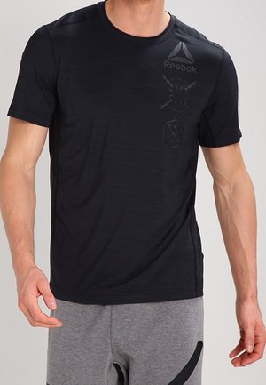 Uomo che indossa una maglietta nera Reebok a maniche corte e pantaloncini sportivi grigi su sfondo semplice.