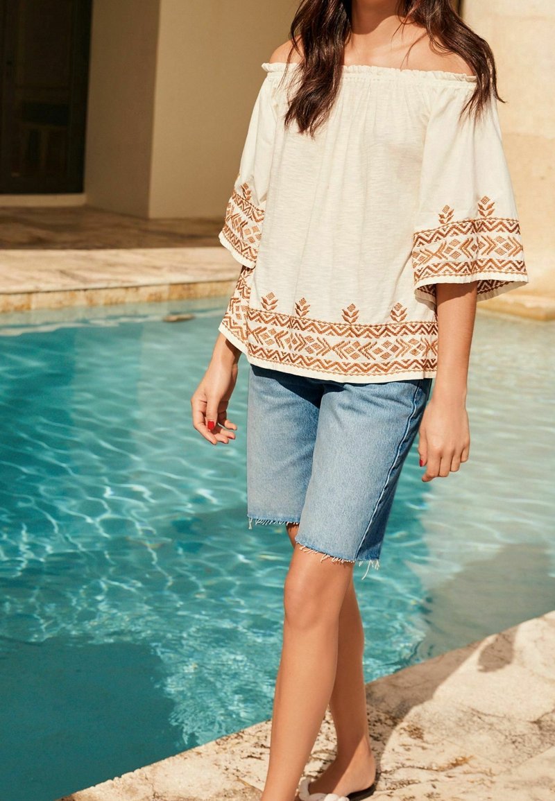 Femme portant un haut blanc épaules dénudées avec broderie marron et un short en jean mi-cuisse, debout au bord de la piscine.