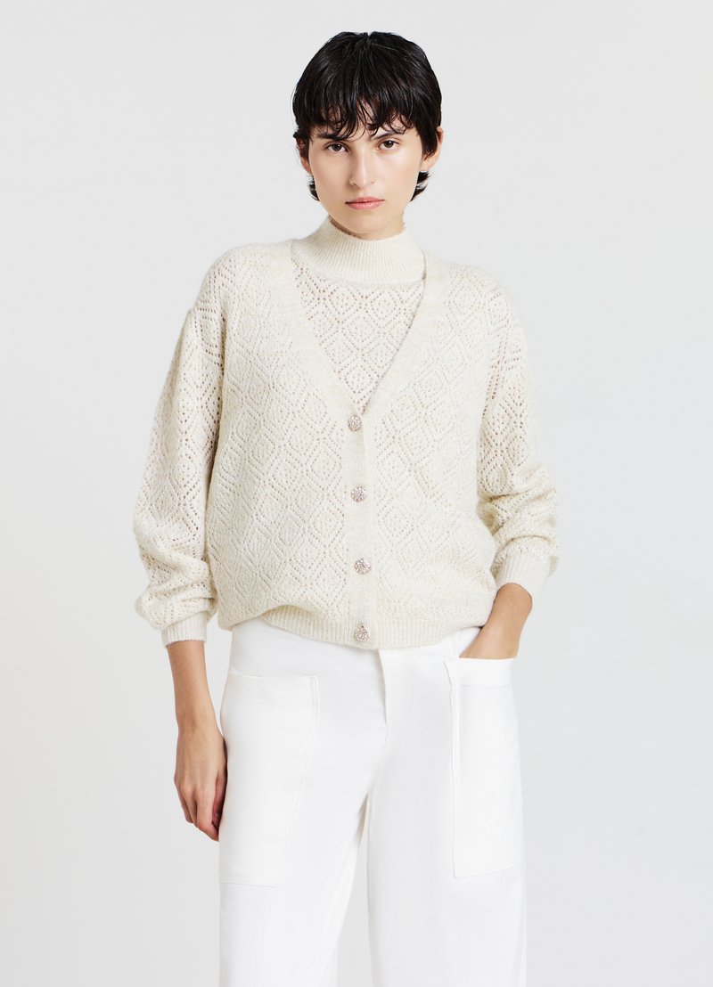 Strickjacke in Creme mit einem Rautenmuster, ausgestattet mit einem tiefen V-Ausschnitt, Knopfverschluss und übergroßen Ärmeln, kombiniert mit einem Rollkragenpullover darunter.