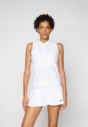 Femme aux cheveux bouclés portant un polo blanc sans manches et une jupe de tennis blanche avec un logo Nike noir, debout devant un fond uni.