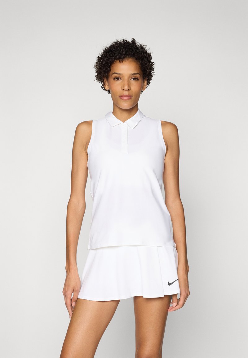 Kobieta z kręconymi włosami, ubrana w białą bezrękawnikową koszulkę polo i białą spódniczkę tenisową z czarnym logo Nike, stojąca na jednolitym tle.