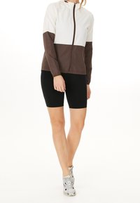 Veste color-block en haut blanc et bas marron foncé, avec une fermeture éclair intégrale et une capuche ; associée à un short noir ajusté.