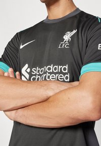 Tummanharmaa jalkapallo-asu turkooseilla yksityiskohdilla, jossa on Liverpool FC:n logo, Niken swoosh ja valkoinen "Standard Chartered" -logo.