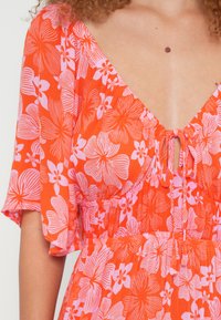 Ziedu kleita spilgti oranžā krāsā ar rozā ziedu rakstiem. Ir dziļš V veida izgriezums un īsas piedurknes ar savilkumu pie jostas.