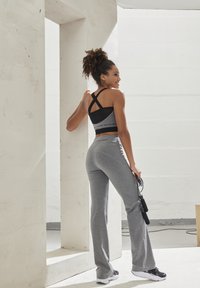 Schwarzer und grauer Sport-BH mit gekreuztem Rücken, kombiniert mit grauen weit ausgestellten Sporthosen. Das Model hält ein Springseil in einem hellen Innenraum.