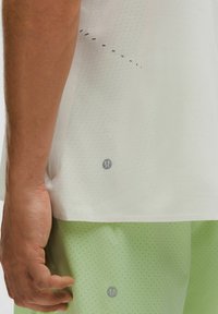 Nærbillede af en hånd ved siden af en hvid perforeret sportsbluse og lysegrønne perforerede shorts med små reflekterende logoer.