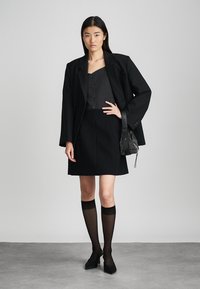 Blazer noir ajusté sur un top et une jupe noirs, associé à des chaussettes noires semi-transparentes jusqu'aux genoux et des escarpins noirs à bout pointu. Petit sac texturé.