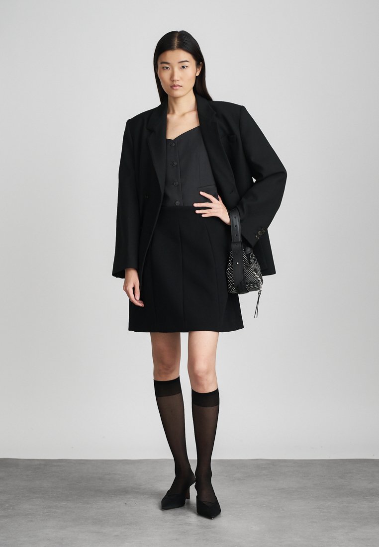 Blazer noir ajusté sur un top et une jupe noirs, associé à des chaussettes noires semi-transparentes jusqu'aux genoux et des escarpins noirs à bout pointu. Petit sac texturé.