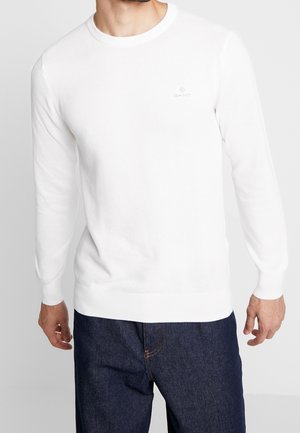 Homme portant un pull blanc texturé à col rond et un jean bleu foncé, debout devant un fond clair uni.
