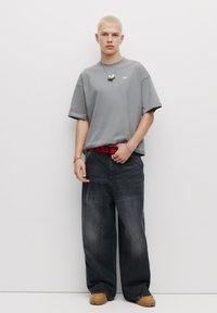 Grå oversized t-shirt, mørke baggy jeans, rød mønstret bælte, beige chunky sko, sølv vedhængs halskæde, poseret foran en hvid baggrund.