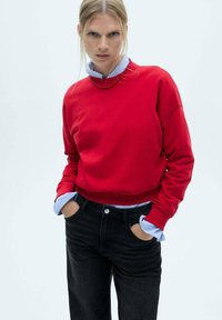 Red cotton sweater with a crew neck, featuring long sleeves, worn over a light blue collared shirt. Paired with black jeans.