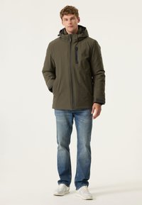 Olijfgroene, geïsoleerde jas met capuchon, rits aan de voorkant en borstzak. Gedragen over blauwe spijkerbroeken en lichtgrijze sneakers.