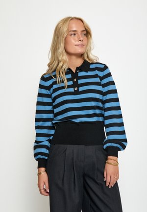 Pull polo en tricot noir et bleu à rayures, avec des manches longues, une patte de boutonnage à trois boutons et un ourlet noir côtelé. Porté avec un pantalon taille haute.