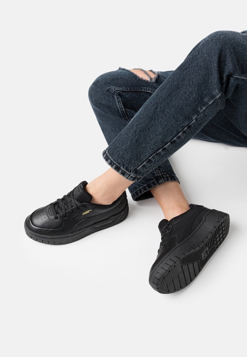 Chaussures de sport noires avec une tige en cuir texturé, une pointe ronde et une semelle en caoutchouc avec un motif de traction. Pas de logos ni d'accents visibles.