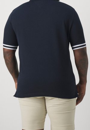Person iført en mørkeblå kortærmet polo med hvide stribedetaljer og beige shorts, vist bagfra.
