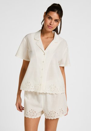 Vrouw die een witte blouse met korte mouwen en knopen draagt en bijpassende korte broek met geschulpte randen en bloemen uitsnijdingen.