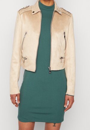 Chaqueta de motorista de ante beige con cremalleras plateadas, usada sobre un vestido ajustado de manga larga, verde oscuro, hasta la rodilla, en una persona de pie.