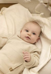 Een beige, textuur rijke lange mouwen shirt gedragen door een baby op een lichtgekleurde deken. Zachte, neutrale tinten met een klein leren accent.