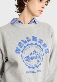 Sudadera gris con un diseño gráfico azul que incluye el texto "WELLNESS" y una ilustración de una montaña, superpuesta a un collar de camisa a rayas azules y blancas.