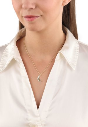 Collana d'oro con ciondolo a forma di luna crescente con superficie testurizzata, indossata da un modello con una camicia di raso bianca.