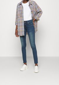 Camisa de cuadros multicolor con dos bolsillos, camiseta blanca debajo, jeans azul oscuro y zapatillas blancas. Diseño estructurado y ajustado.