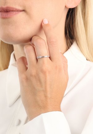 Anello in argento con piccoli motivi a forma di cuore disposti in un pattern, indossato su una mano che tocca il mento, su uno sfondo bianco.