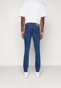 Modré džíny s úzkým střihem, se koženou nášivkou vzadu, pěti kapsami a minimálním detailem šití. Na nohou bílé tenisky.