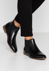 Botas de tobillo de cuero negro con paneles laterales elásticos, punta redondeada y un bajo tacón de madera, llevadas con pantalones ajustados negros.