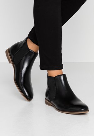 Bottes de cheville en cuir noir avec des panneaux latéraux élastiques, un bout arrondi et un petit talon en bois, portées avec un pantalon noir ajusté.