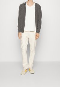 Grauer Kapuzenpullover über einem weißen Hemd, kombiniert mit hellen Hosen und beigen Sneakers mit gelben Akzenten. Einfaches Design, lässiger Schnitt.