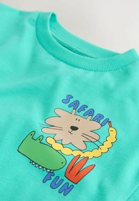 Türkisfarbenes Baumwoll-T-Shirt mit einem verspielten Motiv eines Löwen, Krokodils, einer Schlange und dem Schriftzug "SAFARI FUN" in kräftigen Farben.