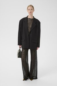 Blazer oversize nero sopra un top scintillante e pantaloni di pizzo nero trasparente. Gli accessori includono una piccola borsa grigia e tacchi a punta verdi.